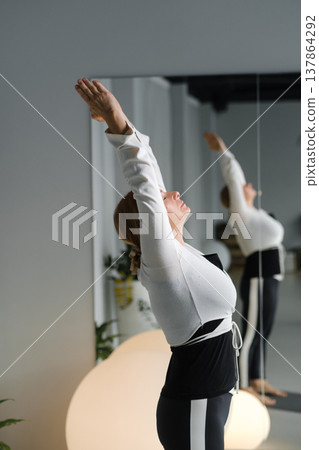 Woman stretches her arms upward in a bright room with mirrors and plants during a wellness session in the afternoon Woman stretches her arms upward in a bright room with mirrors and plants during a wellness session in the afternoon 137864292
