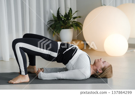 Woman practicing bridge pose on yoga mat in a bright room with plants and soft lighting in the background Woman practicing bridge pose on yoga mat in a bright room with plants and soft lighting in the background 137864299