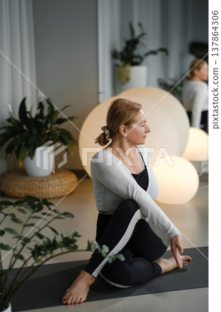 Practicing yoga in a studio with soft lighting and plants while a woman sits on a mat and performs a seated twist at an evening time 137864306
