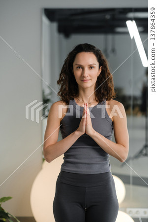 Woman practices yoga in a studio with plants and bright lights during the day while standing in a prayer position with hands together Woman practices yoga in a studio with plants and bright lights during the day while standing in a prayer position with hands together 137864359