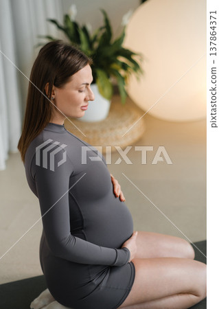 Pregnant woman in yoga pose performing meditation indoors during morning light in a calm space Pregnant woman in yoga pose performing meditation indoors during morning light in a calm space 137864371