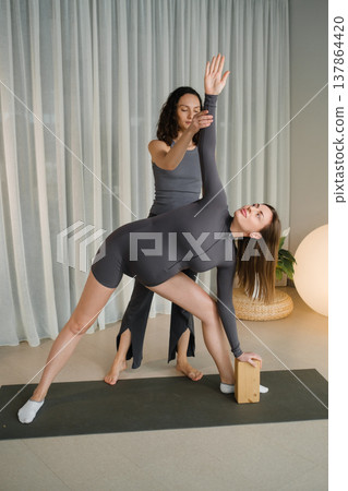 Yoga class takes place in a bright studio where two women practice a pose with support from a teacher and a yoga block Yoga class takes place in a bright studio where two women practice a pose with support from a teacher and a yoga block 137864420