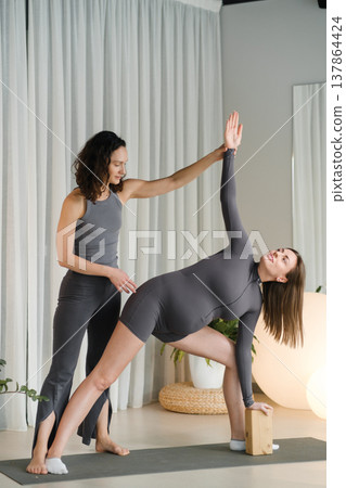 Yoga class takes place in a bright studio where two women practice a pose with support from a teacher and a yoga block Yoga class takes place in a bright studio where two women practice a pose with support from a teacher and a yoga block 137864424