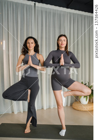 Two women practice yoga indoors in a bright room with white curtains during the day, focusing on balance and connection through their poses and breathing Two women practice yoga indoors in a bright room with white curtains during the day, focusing on balance and connection through their poses and breathing 137864426