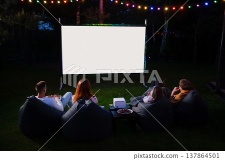 Friends enjoy movie night in backyard under string lights with a projector and bean bags at dusk Friends enjoy movie night in backyard under string lights with a projector and bean bags at dusk 137864501