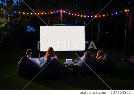 People enjoy outdoor movie night with a projector and bright lights in the backyard during the evening People enjoy outdoor movie night with a projector and bright lights in the backyard during the evening 137864502