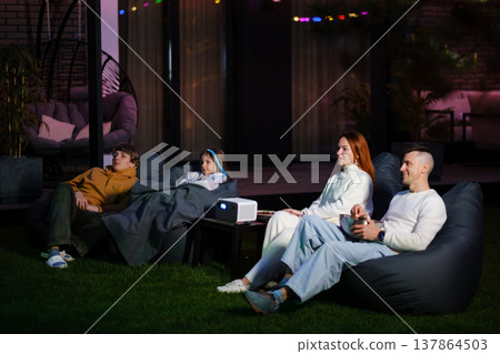 People enjoy movie night outdoors in their backyard using a projector while sitting on bean bag chairs under string lights during the evening People enjoy movie night outdoors in their backyard using a projector while sitting on bean bag chairs under string lights during the evening 137864503