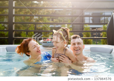 Family enjoys time together in hot tub during afternoon at home while laughing and playing with daughter in warm weather, creating happy moments and memories Family enjoys time together in hot tub during afternoon at home while laughing and playing with daughter in warm weather, creating happy moments and memories 137864570