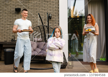Family enjoys a sunny day outside their home while serving food and having fun together in the backyard garden area near a cozy seating area Family enjoys a sunny day outside their home while serving food and having fun together in the backyard garden area near a cozy seating area 137864588
