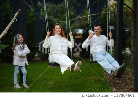 Family enjoys time together swinging in the park during daytime in a backyard with trees and greenery 137864601
