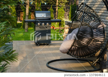 Grill stands on patio next to hanging chair in green backyard during sunny afternoon Grill stands on patio next to hanging chair in green backyard during sunny afternoon 137864611