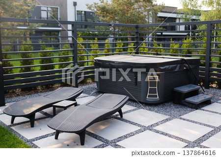 Hot tub area with lounge chairs in a backyard setting on a sunny day in a residential neighborhood surrounded by trees and a fence Hot tub area with lounge chairs in a backyard setting on a sunny day in a residential neighborhood surrounded by trees and a fence 137864616