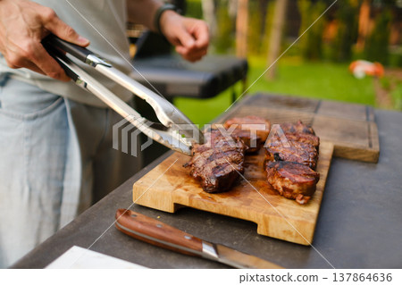 Grilling steak outdoors at a backyard barbecue in warm weather with friends enjoying time together in the evening sunlight Grilling steak outdoors at a backyard barbecue in warm weather with friends enjoying time together in the evening sunlight 137864636
