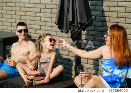Family enjoys summer day by the pool with fruit and drinks while wearing swimwear and sunglasses Family enjoys summer day by the pool with fruit and drinks while wearing swimwear and sunglasses 137864663