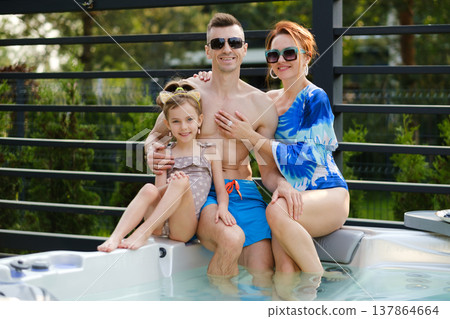 Family enjoys time by a hot tub at a backyard on a sunny day while smiling and relaxing together with fruit on a table nearby Family enjoys time by a hot tub at a backyard on a sunny day while smiling and relaxing together with fruit on a table nearby 137864664