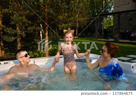 Family enjoys time in a hot tub on a sunny day in a backyard with trees and a house in the background while a girl stands in the water with joy 137864675