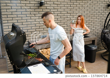 Man cooks food on grill while woman watches and holds drink in outdoor kitchen area during sunny day Man cooks food on grill while woman watches and holds drink in outdoor kitchen area during sunny day 137864714