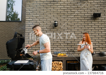 Two people cook food on a barbecue grill at a backyard barbecue while one enjoys a drink on a sunny day in a residential setting Two people cook food on a barbecue grill at a backyard barbecue while one enjoys a drink on a sunny day in a residential setting 137864716