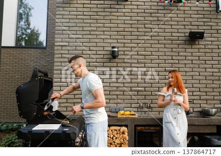 People grilling food outside near a house with light-colored walls during a sunny day with trees and a garden in the background enjoying a casual gathering People grilling food outside near a house with light-colored walls during a sunny day with trees and a garden in the background enjoying a casual gathering 137864717