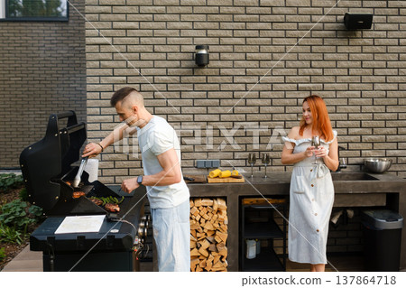People grilling food outside near a house with light-colored walls during a sunny day with trees and a garden in the background enjoying a casual gathering People grilling food outside near a house with light-colored walls during a sunny day with trees and a garden in the background enjoying a casual gathering 137864718