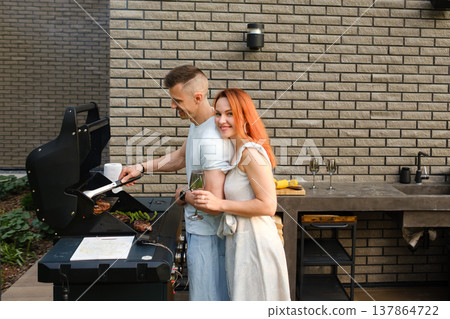 Couple enjoys grilling food outdoors while holding drinks near a modern kitchen in a backyard setting during a sunny day Couple enjoys grilling food outdoors while holding drinks near a modern kitchen in a backyard setting during a sunny day 137864722