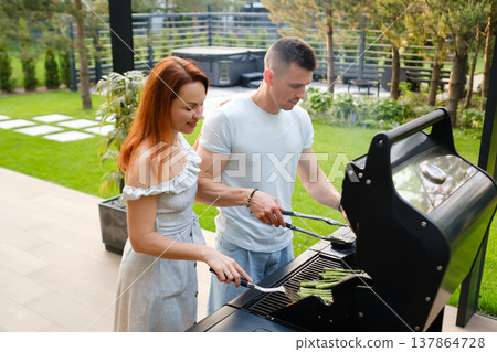 Couple cooking asparagus on grill during summer afternoon in backyard with green plants around and hot tub in background 137864728