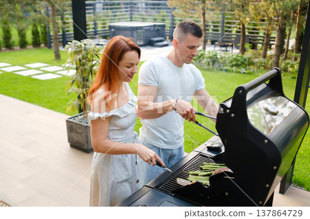People grilling food outdoors on a sunny day in a garden setting with green grass and trees around them in a backyard 137864729