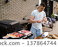 Man prepares meat for grilling at home backyard during afternoon with grill and table set up nearby 137864744