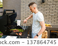 Cooking meat and vegetables on barbecue grill in backyard during sunny afternoon with friends nearby enjoying food and drinks 137864745
