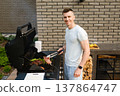 Man stands by a grill preparing meat and vegetables in a backyard setting during daytime with a brick wall as background 137864747