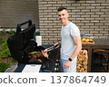 Man cooks meat on a grill in a backyard during a sunny day while smiling at the camera 137864749
