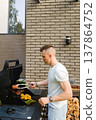 Man cooks meat and vegetables on a grill outside a house during daytime in a backyard 137864752