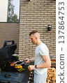 Grilling food outdoors in a backyard during a sunny day with a man preparing meat and vegetables on a barbecue 137864753