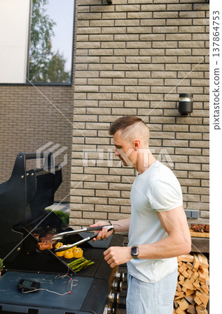 Grilling food outdoors in a backyard during a sunny day with a man preparing meat and vegetables on a barbecue Grilling food outdoors in a backyard during a sunny day with a man preparing meat and vegetables on a barbecue 137864753