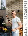 Man grilling meat and vegetables outside near a modern building on a sunny day, enjoying time with friends and family 137864755
