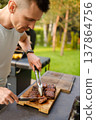 Man slices grilled meat outside in backyard during a sunny day with green trees around and summer vibe in the air 137864756