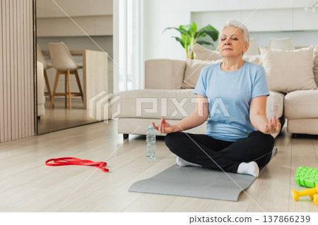 Yoga mindfulness meditation. Senior adult mature woman practicing yoga at home. Mid age old lady sitting in lotus pose on yoga mat meditating relaxing. Older middle aged woman doing breathing practice Yoga mindfulness meditation. Senior adult mature woman practicing yoga at home. Mid age old lady sitting in lotus pose on yoga mat meditating relaxing. Older middle aged woman doing breathing practice 137866239
