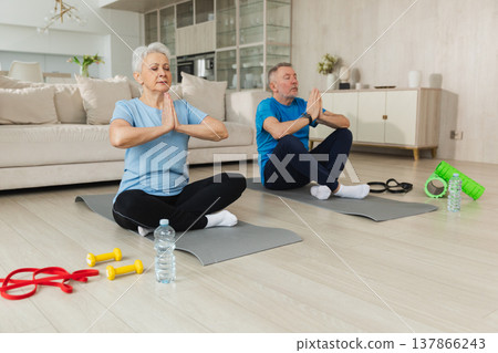 Yoga mindfulness meditation. Senior adult mature couple practicing yoga at home. Mid age old husband wife sitting in lotus pose on yoga mat meditating relaxing. Family doing breathing practice Yoga mindfulness meditation. Senior adult mature couple practicing yoga at home. Mid age old husband wife sitting in lotus pose on yoga mat meditating relaxing. Family doing breathing practice 137866243