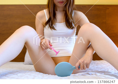 一位年輕女子在臥室裡對著手鏡,用 T 型剃刀刮比基尼部位的毛髮,進行自我照顧。 一位年輕女子在臥室裡對著手鏡,用 T 型剃刀刮比基尼部位的毛髮,進行自我照顧。 137866247