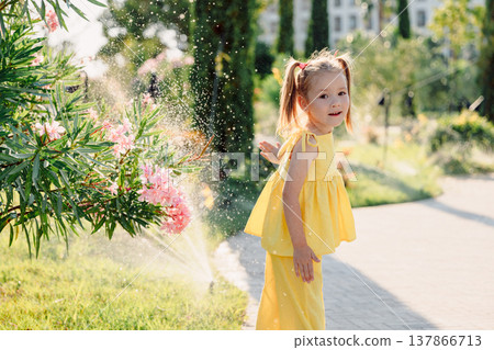 Cute child girl in stylish dress playing near sprinkler on a sunny day in blooming summer park. Cute child girl in stylish dress playing near sprinkler on a sunny day in blooming summer park. 137866713