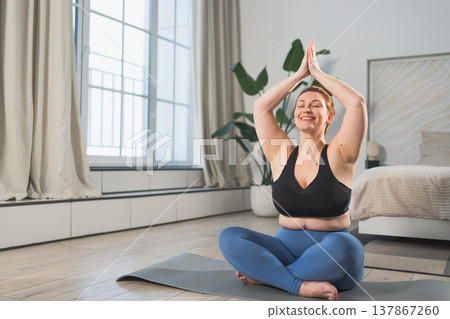 Yoga mindfulness meditation. Woman practicing yoga in living room at home. Plus size female sitting in lotus pose meditating smiling relaxing indoor. Girl doing breathing practice. Yoga at home 137867260