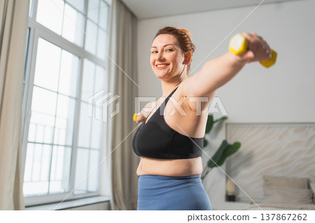 Fitness workout training. Woman picking up dumbbells. Plus size female doing sports exercise at home indoor. Girl in sportswear have workout weight training for weight loss. Sport and fitness Fitness workout training. Woman picking up dumbbells. Plus size female doing sports exercise at home indoor. Girl in sportswear have workout weight training for weight loss. Sport and fitness 137867262