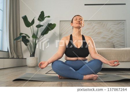 Yoga mindfulness meditation. Woman practicing yoga in living room at home. Plus size female sitting in lotus pose meditating smiling relaxing indoor. Girl doing breathing practice. Yoga at home 137867264