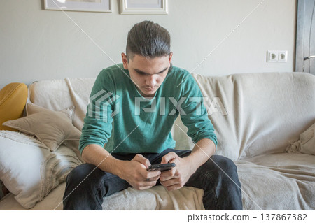A teenager sits on a couch in a living room playing a video game on a mobile phone 137867382