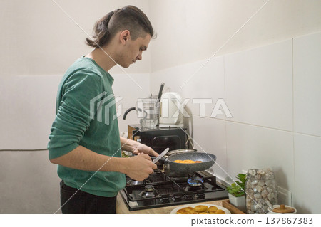 A young man stirs a pot on a stove in a modern kitchen. He is focused on cooking while vegetables  137867383