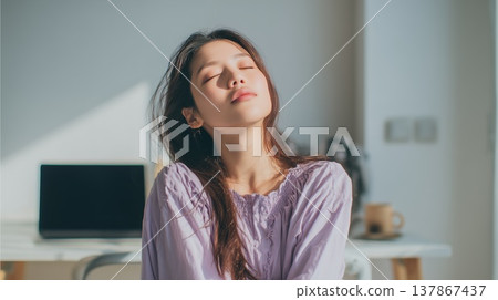 A woman stops working on her computer in a bright room filled with sunlight, closes her eyes, and rests, basking in the sun. 137867437