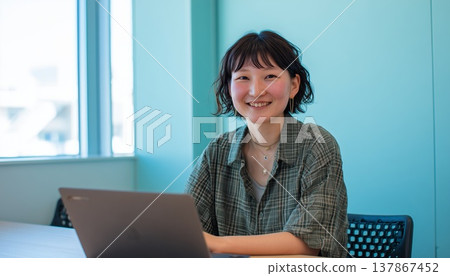 白天，一位年輕女子在一間牆壁呈現獨特藍色的辦公室裡使用筆記型電腦工作，臉上洋溢著燦爛的笑容。 137867452