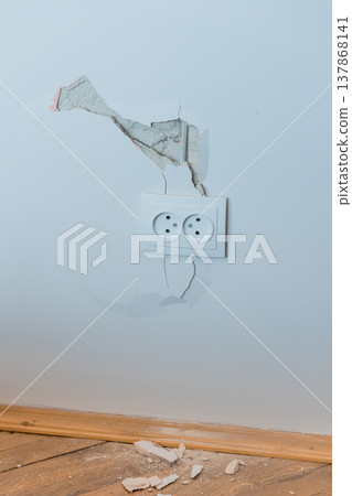Damaged White Wall With Cracked Plaster Around Electrical Outlet Showing Exposed Concrete And Broken Pieces On Wooden Floor In Light Blue Room 137868141