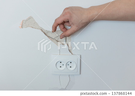 Hand Peeling Cracked White Wall Paint Above Electrical Outlet Showing Damage Repair Needs Realistic Home Maintenance Scene With Neutral Background Texture Detail Hand Peeling Cracked White Wall Paint Above Electrical Outlet Showing Damage Repair Needs Realistic Home Maintenance Scene With Neutral Background Texture Detail 137868144