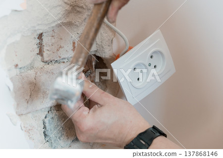Electrician Hammering Wall Near White Dual Socket Outlet During Renovation Project Showing Exposed Brick Texture And Construction Tools In Action Electrician Hammering Wall Near White Dual Socket Outlet During Renovation Project Showing Exposed Brick Texture And Construction Tools In Action 137868146